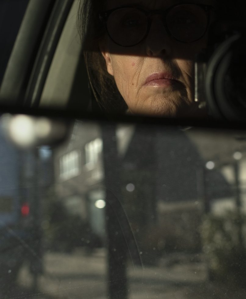 A photograph of a woman driving seen through the rearview mirror of a car.
