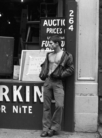 A black-and-white photo of a young man standing on a city sidewalk wearing a jacket with no shirt underneath.