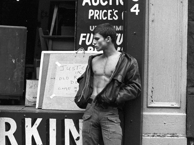 A black-and-white photo of a young man standing on a city sidewalk wearing a jacket with no shirt underneath.