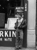 A black-and-white photo of a young man standing on a city sidewalk wearing a jacket with no shirt underneath.