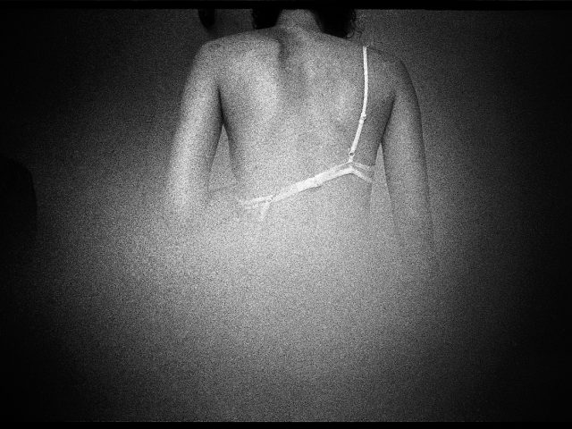 A black and white photograph of a woman in a bra taken from behind.