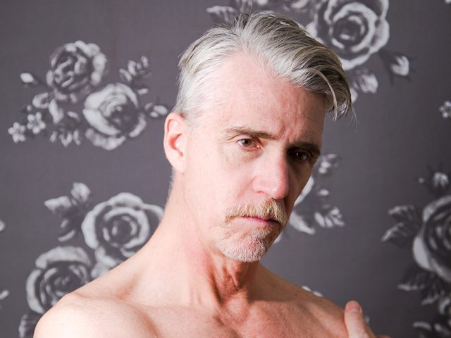 A color photograph of a shirtless man with silver hair.