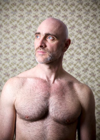 A color photograph os a shirtless man in front of floral wallpaper.