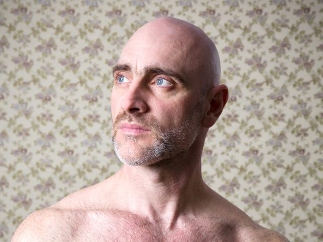 A color photograph os a shirtless man in front of floral wallpaper.