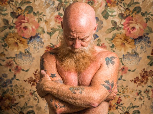 A color photograph of a nude man crossing his arms and looking down.