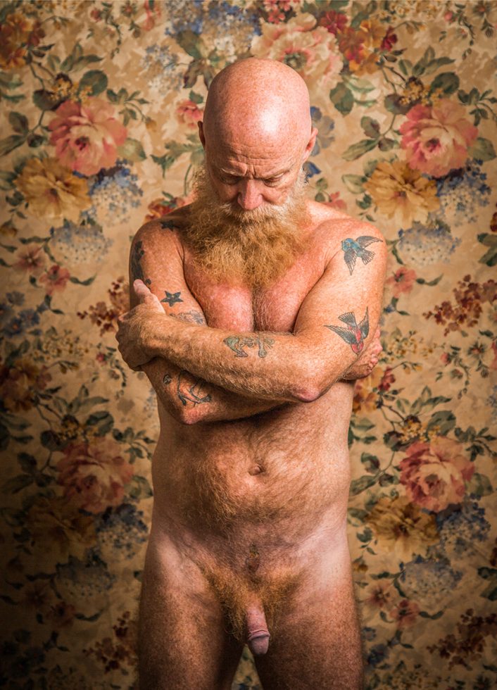 A color photograph of a nude man crossing his arms and looking down.