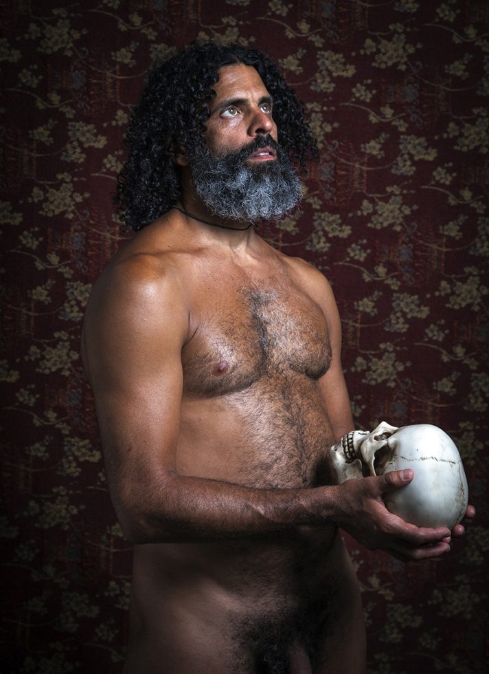 A color photograph of a nude man holding a human skull.