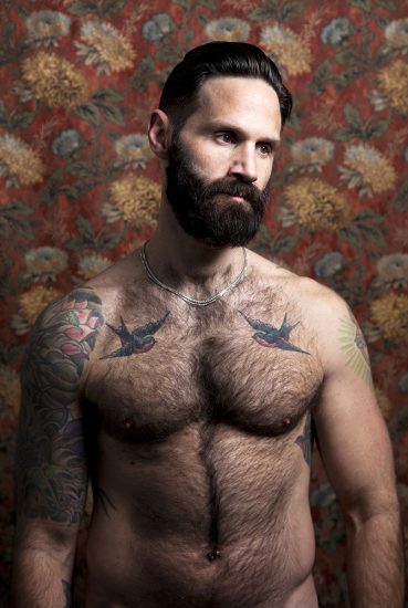 A color photograph os a shirtless hairy man with tattoos.