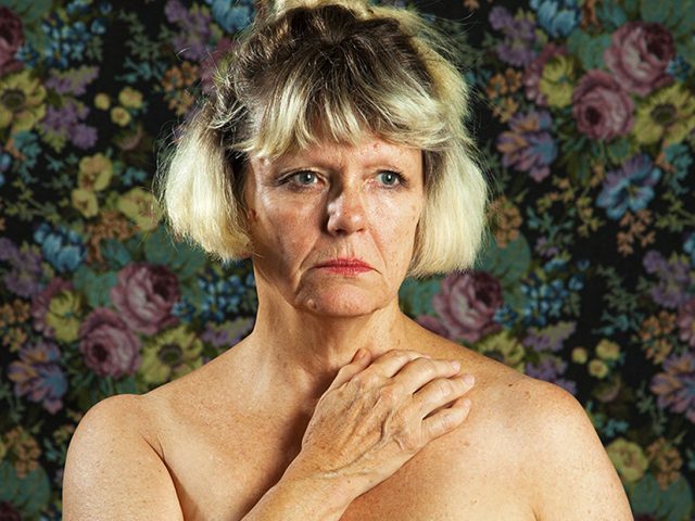 A color photograph os a shirtless woman with her arm across her chest.