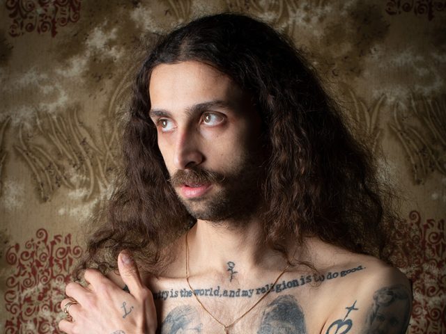 A color photograph of a shirtless man with long hair.