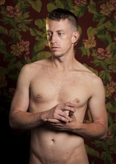 A color photograph os a shirtless man in front of floral wallpaper.
