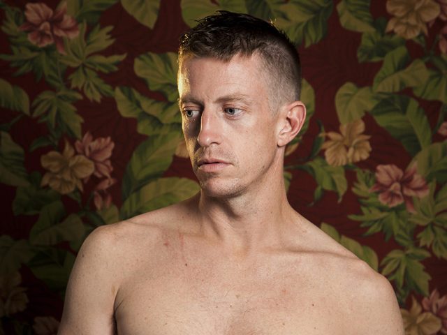 A color photograph os a shirtless man in front of floral wallpaper.