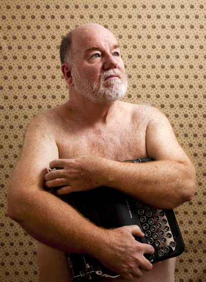 A color photograph of a shirtless man holding a typewriter.