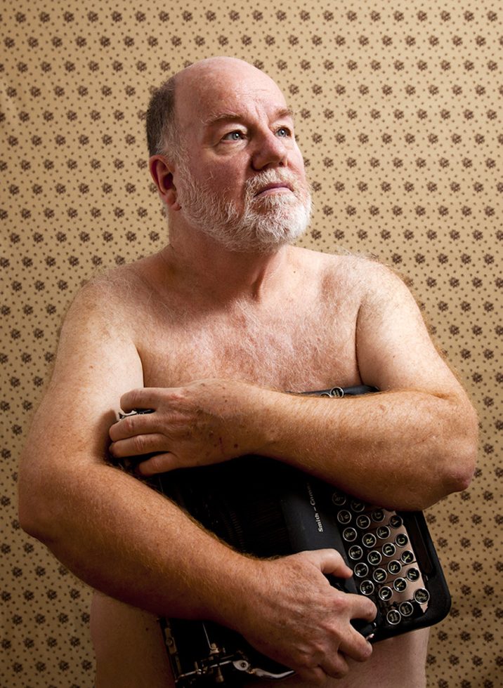 A color photograph of a shirtless man holding a typewriter.