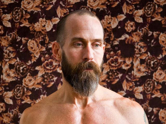 A color photograph of a shirtless man with a beard.
