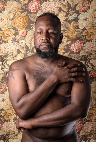 A color photograph of a shirtless man in front of floral wallpaper.
