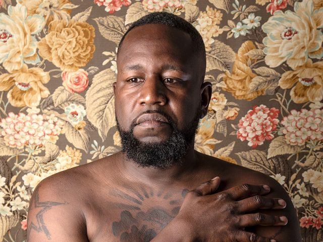 A color photograph of a shirtless man in front of floral wallpaper.