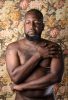 A color photograph of a shirtless man in front of floral wallpaper.