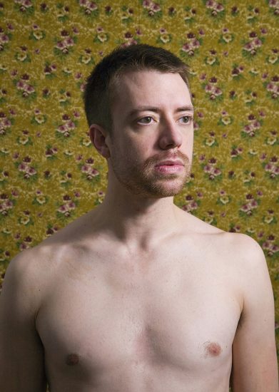 A color photograph of a shirtless man in front of floral wallpaper.