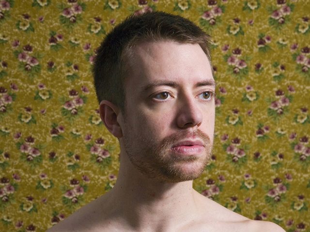 A color photograph of a shirtless man in front of floral wallpaper.