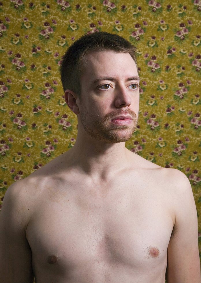 A color photograph of a shirtless man in front of floral wallpaper.