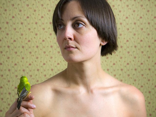 A color photograph os a shirtless woman with a bird perched on her finger.