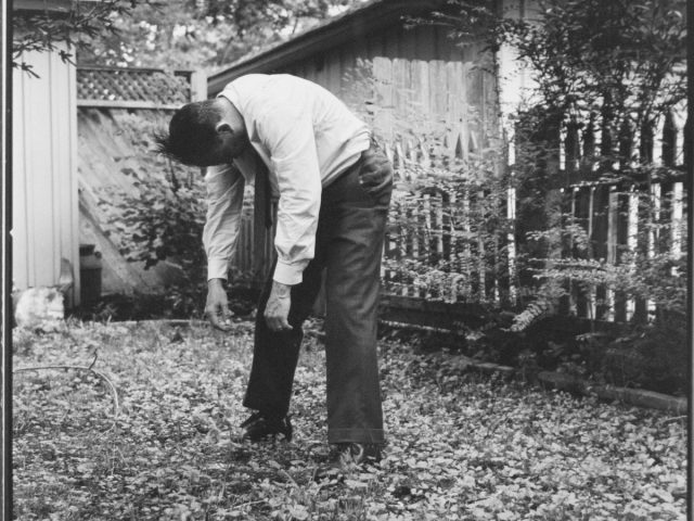 A black and white photograph of a man bent forward in a yard.