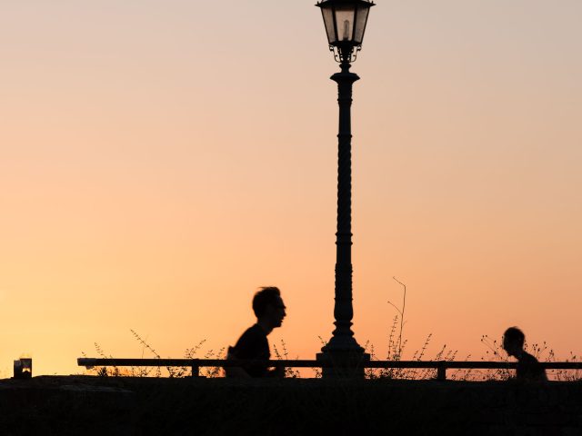 This is a color photograph of two people walking past a lamp post at sunset.