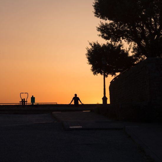 This is a color photograph of a figure sitting and leaning back at a park as the sun sets.