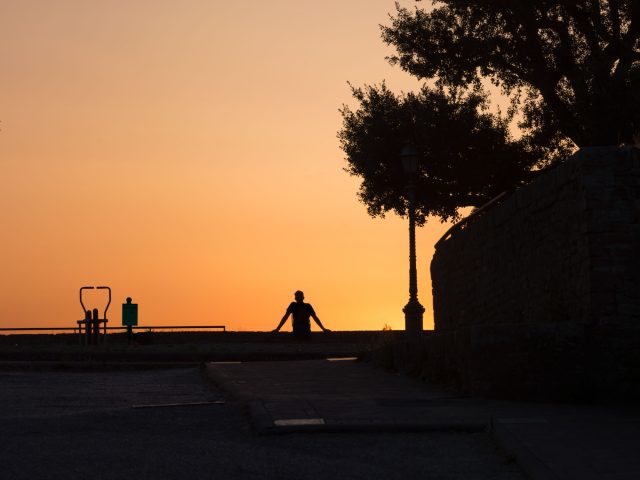 This is a color photograph of a figure sitting and leaning back at a park as the sun sets.