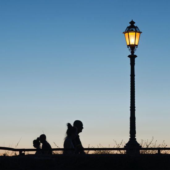 This is a color photograph of a lamp post and people walking as the sun sets.
