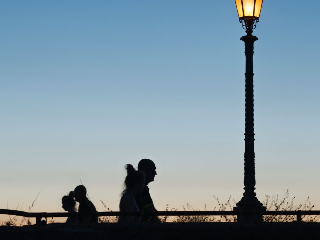 This is a color photograph of a lamp post and people walking as the sun sets.