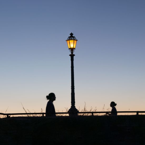 This is a color photograph of a lamp post and two people walking towards each other at dusk.