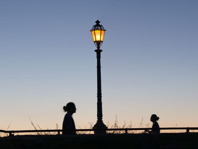 This is a color photograph of a lamp post and two people walking towards each other at dusk.