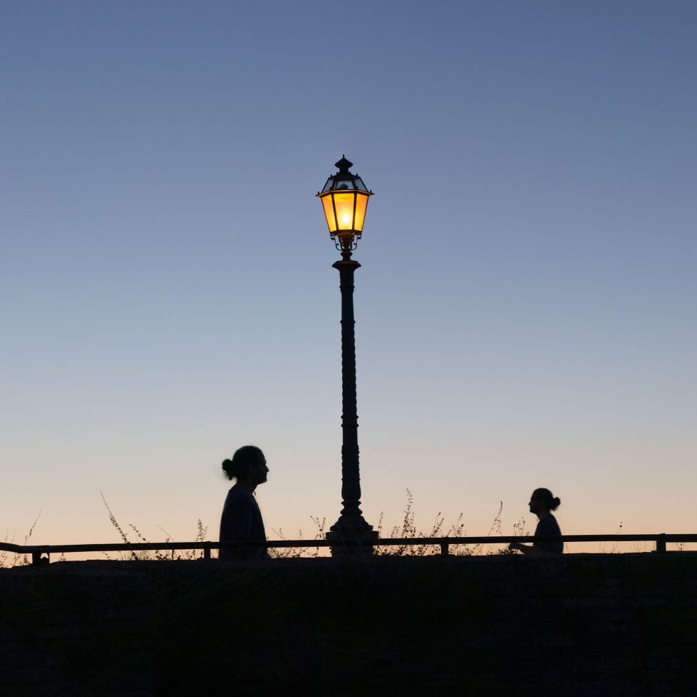 This is a color photograph of a lamp post and two people walking towards each other at dusk.