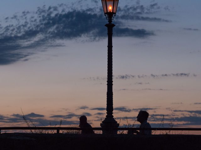 This is a color photograph of two people walking past a lamp post at dusk.