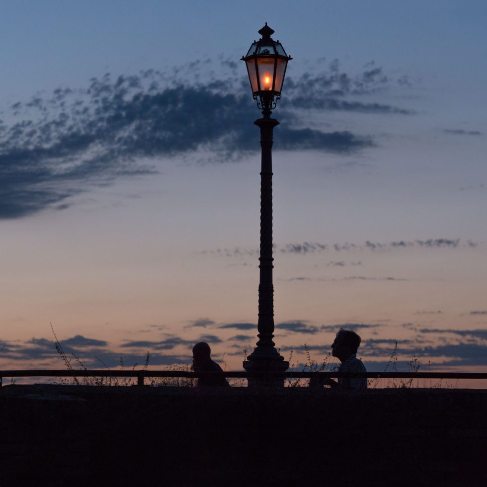 This is a color photograph of two people walking past a lamp post at dusk.