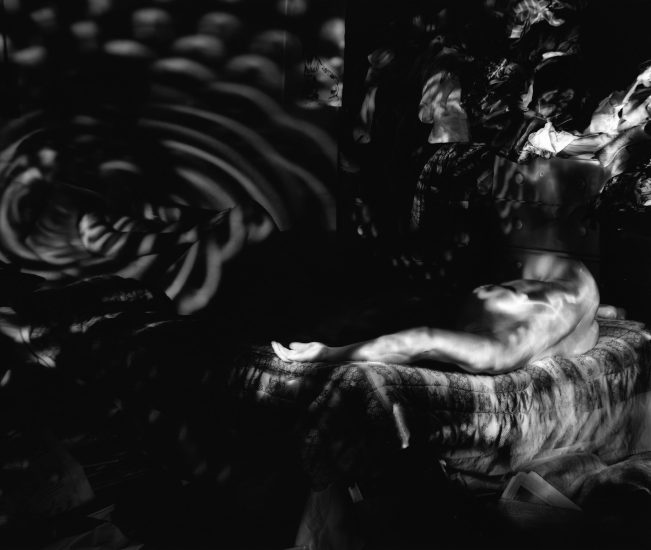 Tis black and white photograph shows ghostly figures sprawled across a bed. Circles of light are cast on the bedroom walls.