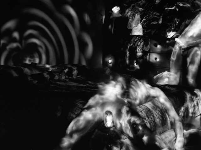 This black and white photo shows a naked man lying relaxed in a chair in his apartment. Strange patterns of light are cast behind him on the wall.
