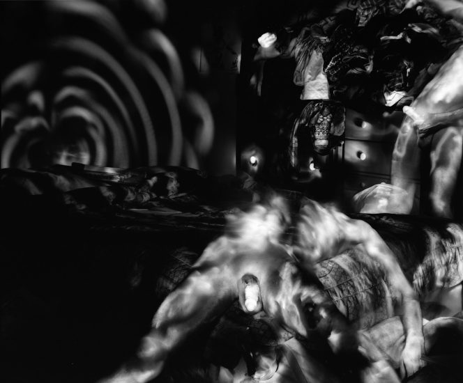 This black and white photo shows a naked man lying relaxed in a chair in his apartment. Strange patterns of light are cast behind him on the wall.