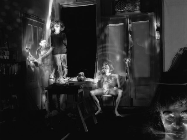 This black and white photograph shows a naked woman, blurred with motion, sitting on a chair in a living room. Other figures surrounding her are blurred with motion.