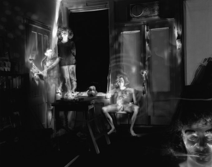 This black and white photograph shows a naked woman, blurred with motion, sitting on a chair in a living room. Other figures surrounding her are blurred with motion.