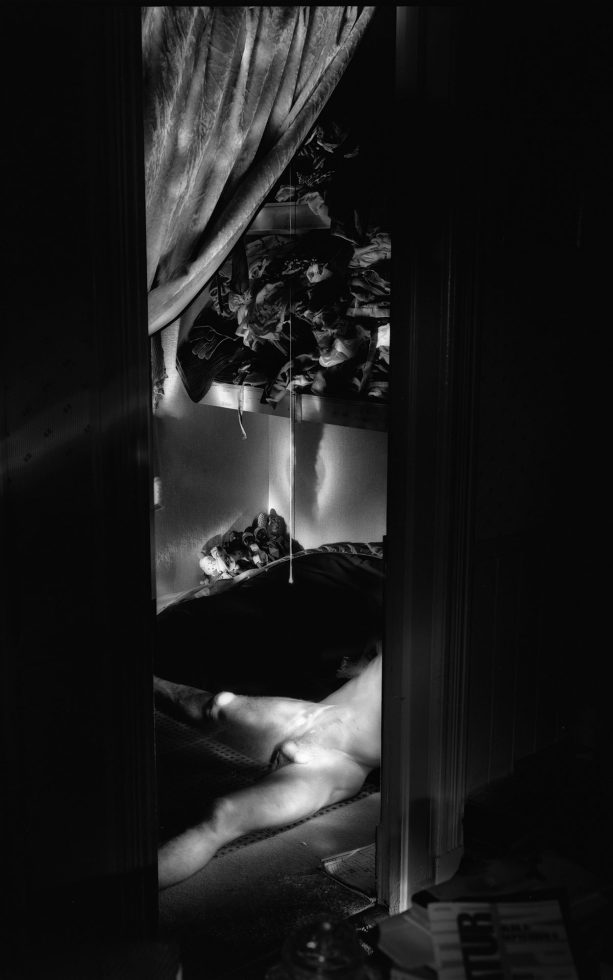 This photograph shows a vertical space seen through a doorway. The naked body of a man is partially visible. He is seen lying on the ground.
