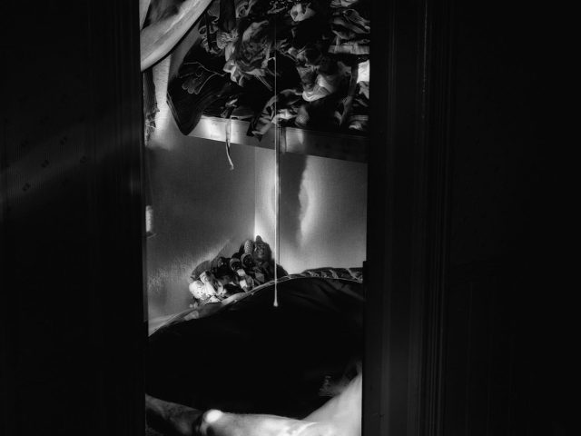 This photograph shows a vertical space seen through a doorway. The naked body of a man is partially visible. He is seen lying on the ground.