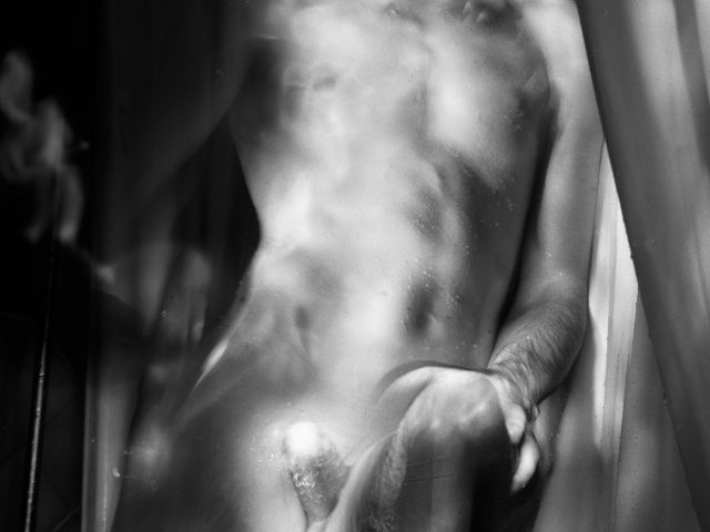 This black and white photograph shows the naked body of a man, seen from the waist up to his neck. He stands in front of curtains.