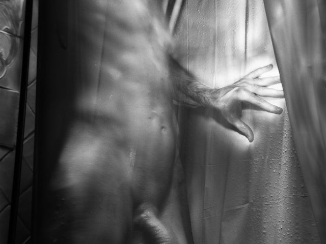 This photograph shows a naked man in a tight space. His hand is glowing and pushing against a curtain.
