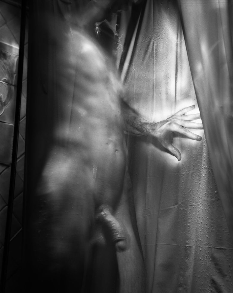 This photograph shows a naked man in a tight space. His hand is glowing and pushing against a curtain.