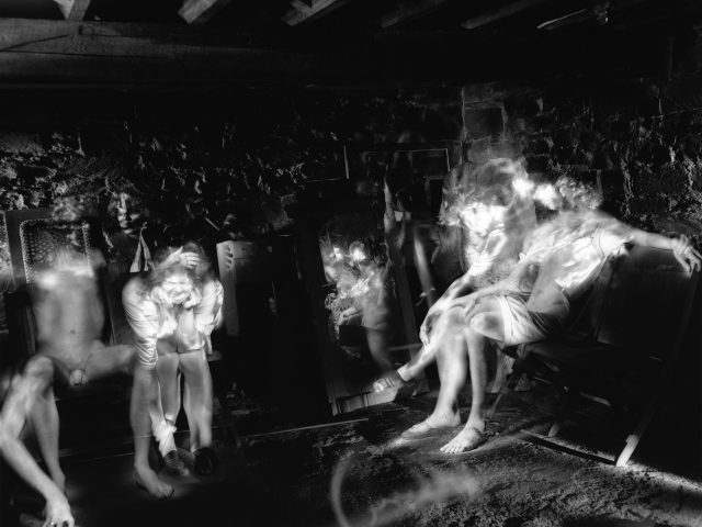 This photograph shows the illuminated figures of naked men, sitting inside a dark basement space.