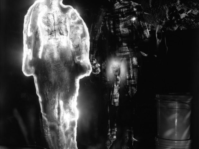 This photograph shows a human figure surrounded by a glowing white line of light. Behind him, the crowded space of a basement is slightly illuminated.