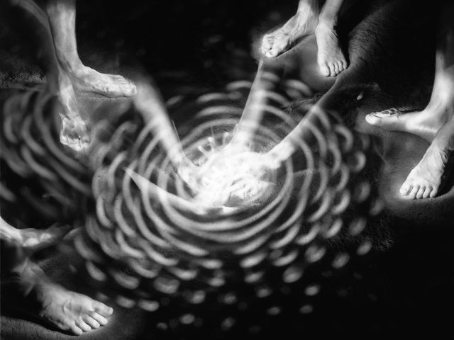 This black and white photograph shows a circle of bare feet glowing against a black background. A strange circle of light is cast on the ground between them.